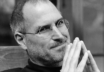 Image is a black and white photo of Steve Jobs, tenting his fingers and smiling, tight-lipped, at the camera.